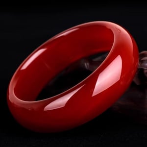 Puede incluir: Una pulsera brazalete de color rojo vibrante. La pulsera pulida y translúcida es una banda sólida de color, que refleja la luz. La joya se encuentra sobre un fondo oscuro, resaltando su rico tono y superficie lisa. Un accesorio clásico.