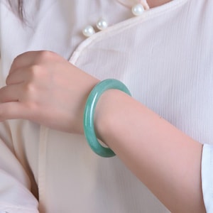 May include: A jade bangle bracelet in a light green hue. The smooth, polished surface reflects light, showcasing the natural variations in color. The bracelet is a solid, circular design, worn on a wrist.