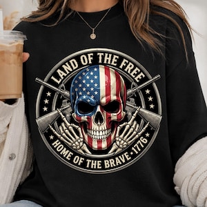 Könnte beinhalten: Schwarzes Sweatshirt mit einem Totenkopf-Design in den Farben der amerikanischen Flagge und gekreuzten Gewehren. Der Text lautet "LAND OF THE FREE, HOME OF THE BRAVE - 1776". Das Design befindet sich auf der Vorderseite.