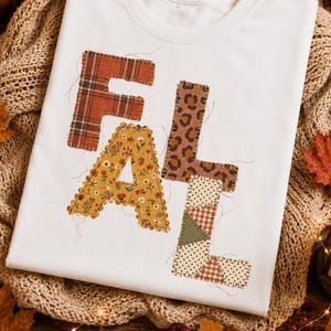 May include: A white t-shirt featuring the word FALL in large, block-style letters. Each letter has a distinct pattern: the F is red plaid, the A is yellow with small flowers, the L is brown leopard print, and the L is a patchwork of dots and plaid.