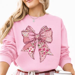 May include: Pink sweatshirt featuring a large bow design. The bow is composed of various pink patterned fabrics, including floral, polka dot, and gingham. The sweatshirt is a solid light pink color.