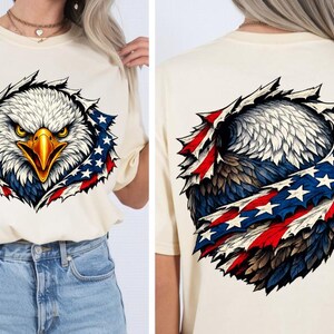May include: A cream-coloured t-shirt with a graphic of an American bald eagle. The eagle's head and chest are visible, surrounded by a design incorporating the American flag's red, white, and blue colours and stars. The back of the shirt also features the eagle design.