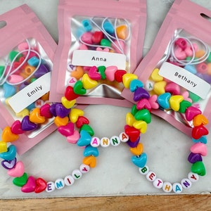May include: Three clear plastic bags with colorful heart-shaped beads and letter beads to make bracelets. The bags are labeled "Emily", "Anna", and "Bethany". The bracelets are made with the beads and spell out the names.