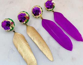 May include: A pair of dangle earrings with green and purple sequin balls and long feathers. One earring has a gold feather and the other has a purple feather.