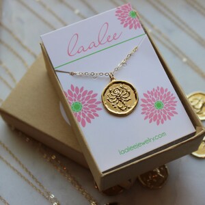 Pu&ograve; includere: Una collana d'oro con un ciondolo rotondo che presenta un design a forma di fiore di loto. La collana &egrave; confezionata in una scatola marrone con un design floreale rosa e verde e il testo "laalee" e "laaleejewelry.com".