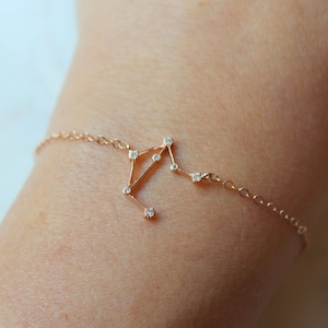 K&ouml;nnte beinhalten: Ein zartes goldenes Armband mit einem Konstellationsdesign, das mit kleinen, klaren Steinen besetzt ist. Die Konstellation ist auf einem Kettenarmband angebracht.