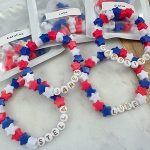 May include: Four bracelets made with red, white, and blue star-shaped beads. The bracelets spell out the names "Stella", "Luke", "James", and "Caroline" in gold letters.