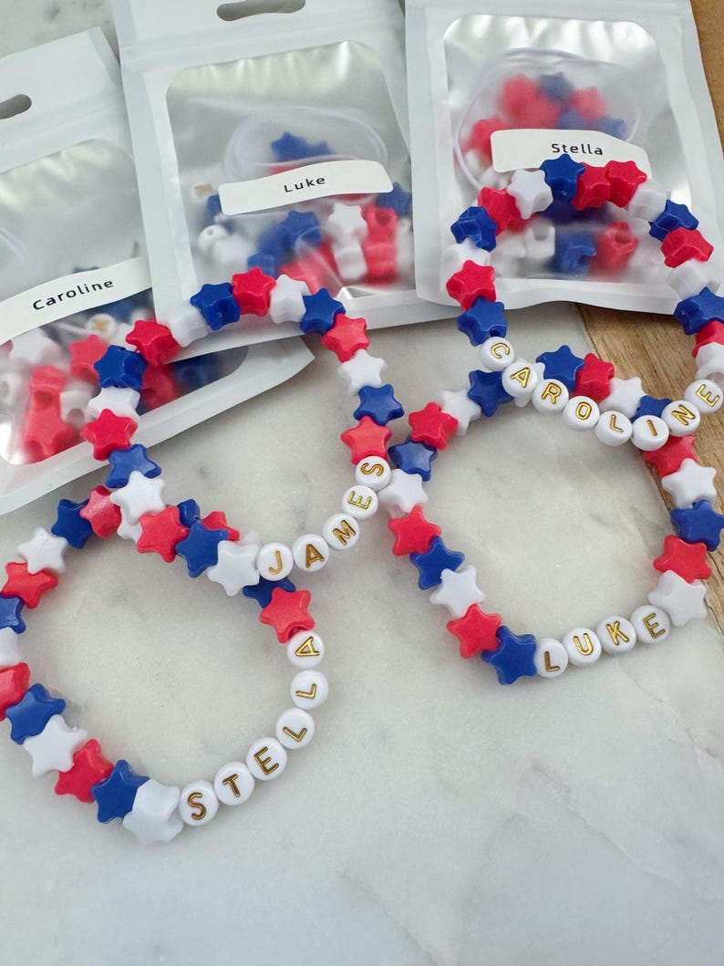 May include: Four bracelets made with red, white, and blue star-shaped beads. The bracelets spell out the names "Stella", "Luke", "James", and "Caroline" in gold letters.