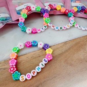 May include: Colorful flower bead bracelets with names spelled out in letter beads. The bracelets feature a variety of flower colors, including pink, orange, green, blue, and purple. The names "Sophie", "Mya", and "Charlotte" are visible.