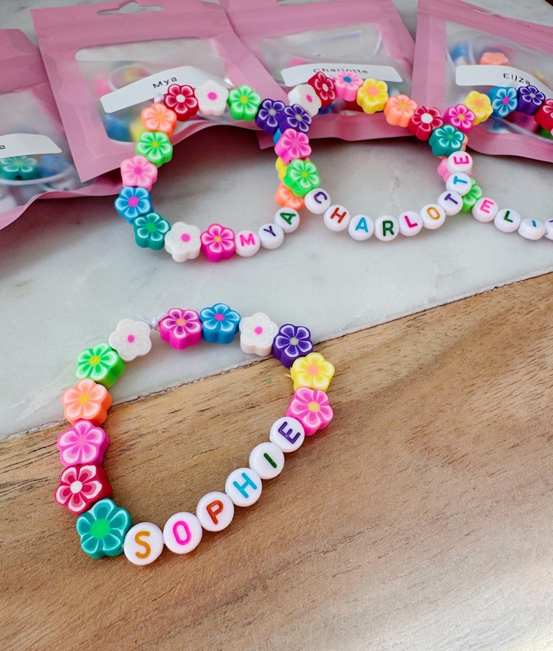 May include: Colorful flower bead bracelets with names spelled out in letter beads. The bracelets feature a variety of flower colors, including pink, orange, green, blue, and purple. The names "Sophie", "Mya", and "Charlotte" are visible.
