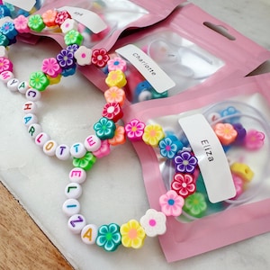May include: Colorful beaded bracelets with flower accents and personalized names. The bracelets feature white letter beads spelling out names, interspersed with vibrant flower-shaped beads in various colors. Packaged in pink pouches.