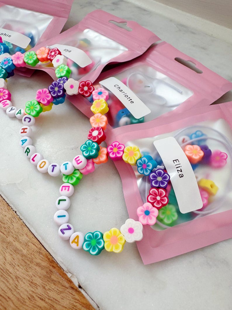 May include: Colorful beaded bracelets with flower accents and personalized names. The bracelets feature white letter beads spelling out names, interspersed with vibrant flower-shaped beads in various colors. Packaged in pink pouches.