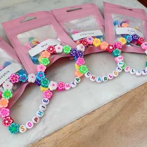 May include: Four personalized bracelets with colorful flower beads and letter beads spelling out names. Each bracelet is packaged in a pink bag. The names on the bracelets are Sophie, Mya, Charlotte, and Eliza.