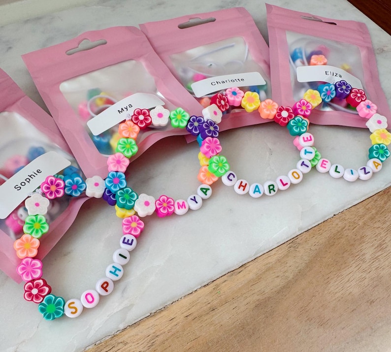 May include: Four personalized bracelets with colorful flower beads and letter beads spelling out names. Each bracelet is packaged in a pink bag. The names on the bracelets are Sophie, Mya, Charlotte, and Eliza.