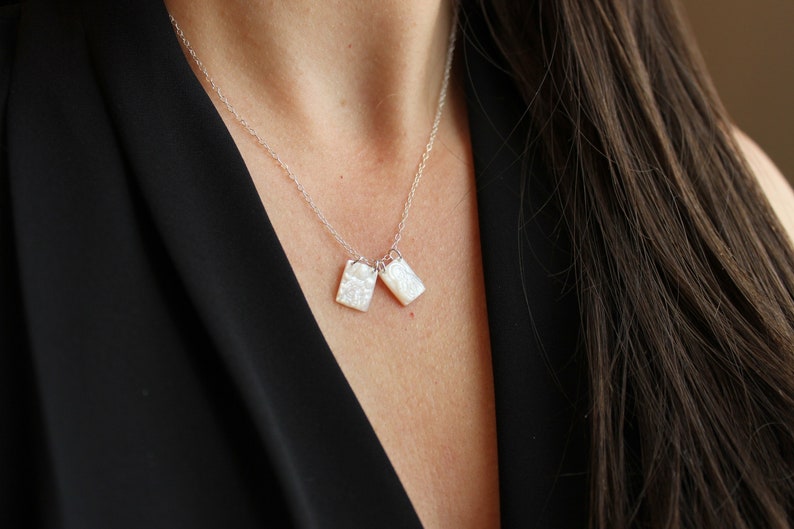 May include: A silver chain necklace with two white rectangular pendants. The pendants have a textured, iridescent surface.