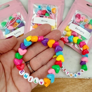 May include: Three small pink bags with colorful heart-shaped beads and letter beads. The bags are labeled "Emily", "Anna", and "Bethany". A rainbow heart-shaped bracelet with the name "Emily" spelled out in white letters is shown in the center of the image.