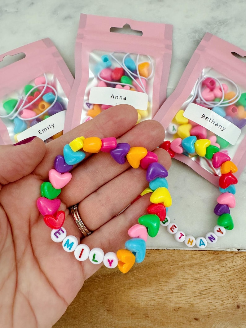 May include: Three small pink bags with colorful heart-shaped beads and letter beads. The bags are labeled "Emily", "Anna", and "Bethany". A rainbow heart-shaped bracelet with the name "Emily" spelled out in white letters is shown in the center of the image.