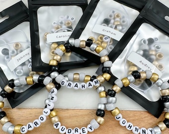 Puede incluir: Cuatro bolsas de plástico transparente con bordes negros contienen cuentas de color negro, blanco y dorado. Las bolsas están etiquetadas con los nombres "Johnny", "Carolyn", "Graham" y "Audrey". Cuatro pulseras hechas con las cuentas deletrean los nombres "Johnny", "Carolyn", "Graham" y "Audrey".