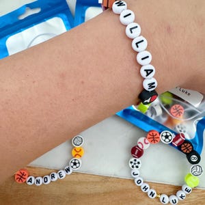 May include: Three bracelets with white beads and black lettering spelling out the names "William", "Andrew", and "John Luke". Each bracelet has colorful sport-themed beads with images of basketballs, baseballs, soccer balls, footballs, and volleyballs.