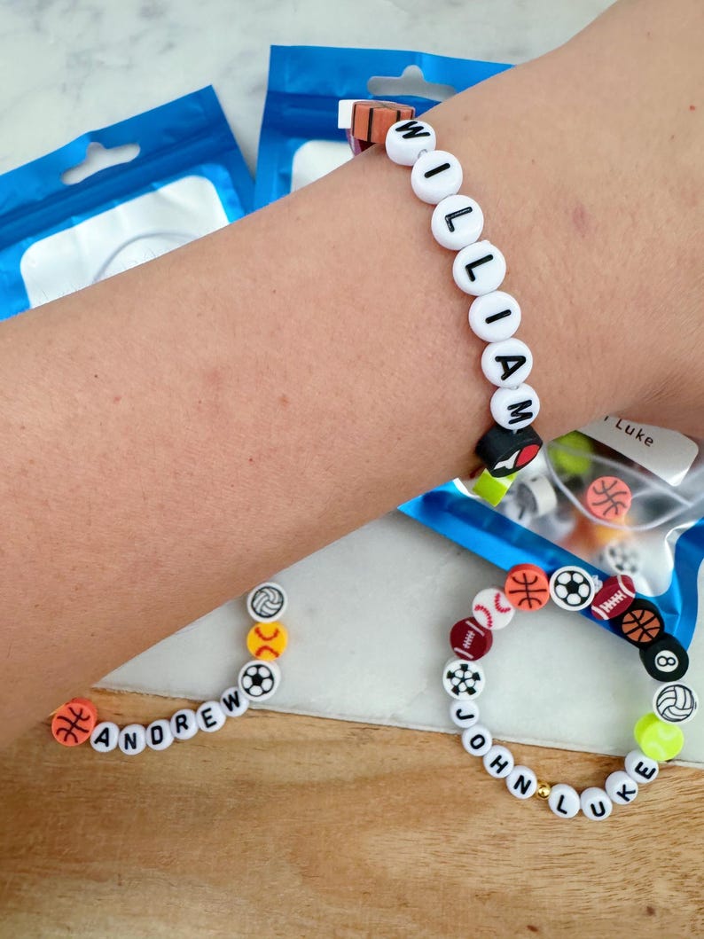 May include: Three bracelets with white beads and black lettering spelling out the names "William", "Andrew", and "John Luke". Each bracelet has colorful sport-themed beads with images of basketballs, baseballs, soccer balls, footballs, and volleyballs.