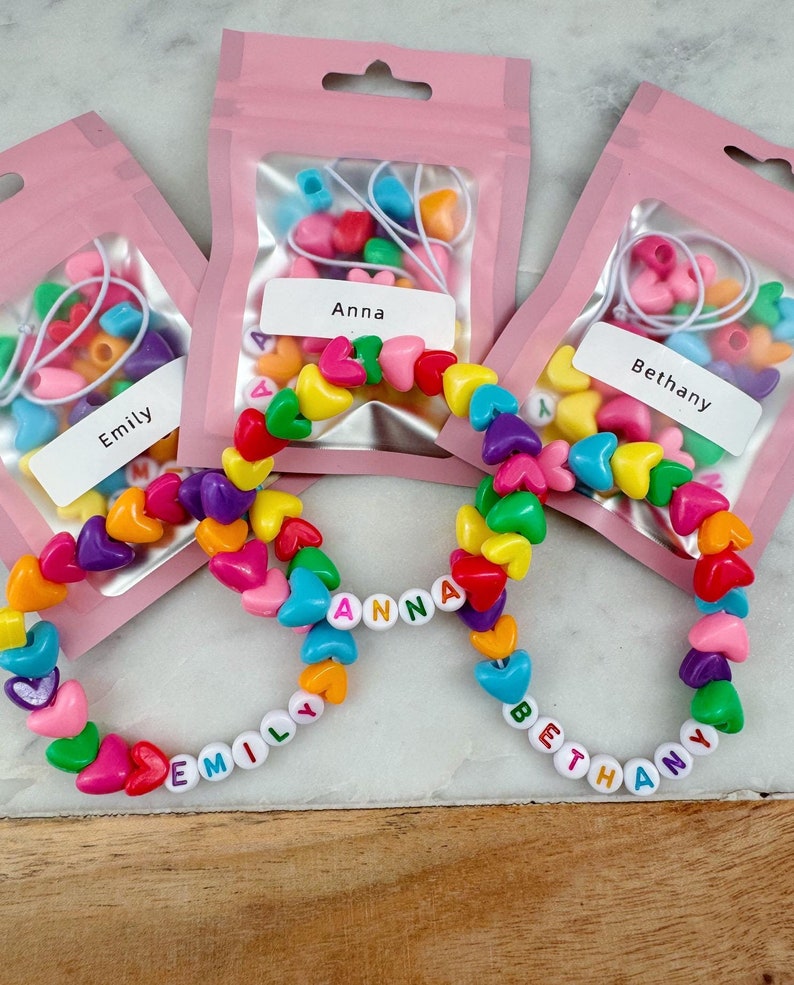May include: Three clear plastic bags with colorful heart-shaped beads and letter beads to make bracelets. The bags are labeled "Emily", "Anna", and "Bethany". The bracelets are made with the beads and spell out the names.