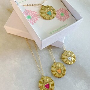 K&ouml;nnte beinhalten: Drei goldene Halsketten mit herzf&ouml;rmigen Anh&auml;ngern. Die Anh&auml;nger sind mit unterschiedlich farbigem Emaille verziert: T&uuml;rkis, Orange und Pink. Die Halsketten werden in einer wei&szlig;en Schachtel mit dem Markennamen "laalee" auf dem Deckel pr&auml;sentiert.