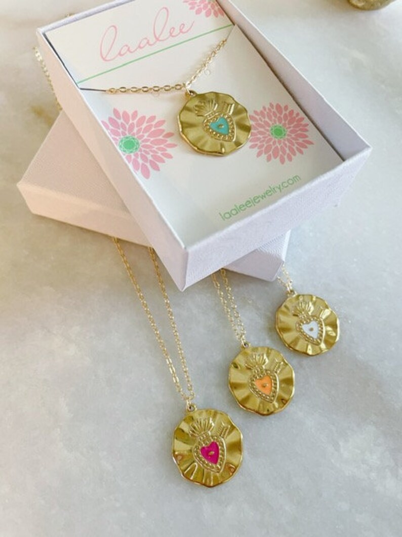 K&ouml;nnte beinhalten: Drei goldene Halsketten mit herzf&ouml;rmigen Anh&auml;ngern. Die Anh&auml;nger sind mit unterschiedlich farbigem Emaille verziert: T&uuml;rkis, Orange und Pink. Die Halsketten werden in einer wei&szlig;en Schachtel mit dem Markennamen "laalee" auf dem Deckel pr&auml;sentiert.