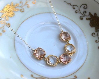 May include: A delicate gold necklace featuring five square-cut gemstones in shades of peach and clear. The gemstones are set in gold bezels and linked by a fine gold chain. The necklace rests on a white plate with gold floral detailing.