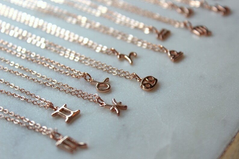 May include: A set of 12 gold-toned zodiac necklaces with delicate chains. Each necklace features a different zodiac symbol, including Aries, Taurus, Gemini, Cancer, Leo, Virgo, Libra, Scorpio, Sagittarius, Capricorn, Aquarius, and Pisces.
