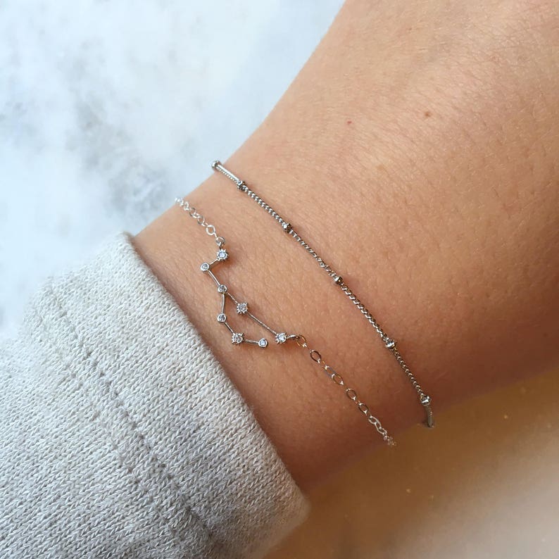 May include: Two silver bracelets on a wrist. The top bracelet is a delicate chain with small beads. The bottom bracelet features a constellation design with small clear stones.