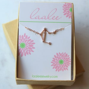 K&ouml;nnte beinhalten: Eine Halskette aus Ros&eacute;gold mit einem zarten Librazier-Anh&auml;nger, der mit kleinen, klaren Zirkonia-Steinen besetzt ist. Die Halskette wird in einer wei&szlig;en Schachtel mit einem rosafarbenen Blumenmuster und dem Text "laalee" und "laaleejewelry.com" pr&auml;sentiert.