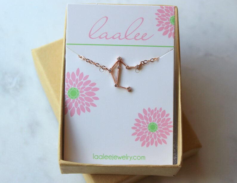 K&ouml;nnte beinhalten: Eine Halskette aus Ros&eacute;gold mit einem zarten Librazier-Anh&auml;nger, der mit kleinen, klaren Zirkonia-Steinen besetzt ist. Die Halskette wird in einer wei&szlig;en Schachtel mit einem rosafarbenen Blumenmuster und dem Text "laalee" und "laaleejewelry.com" pr&auml;sentiert.