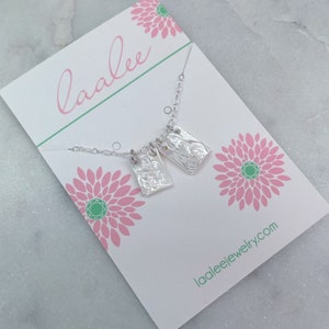 May include: A silver necklace with two rectangular pendants featuring a textured design. The necklace is displayed on a white card with pink flowers and the text "laalee" and "laaleejewelry.com".