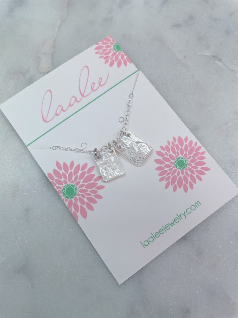 May include: A silver necklace with two rectangular pendants featuring a textured design. The necklace is displayed on a white card with pink flowers and the text "laalee" and "laaleejewelry.com".
