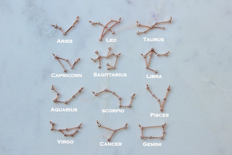 May include: A set of 12 gold zodiac constellation necklaces with small diamonds. The necklaces are arranged in two rows of six, with each necklace labeled with its corresponding zodiac sign: Aries, Taurus, Gemini, Cancer, Leo, Virgo, Libra, Scorpio, Sagittarius, Capricorn, Aquarius, and Pisces.