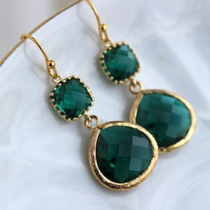 May include: Gold dangle earrings with two emerald green faceted gemstones. The larger gemstone is teardrop shaped and the smaller gemstone is square.