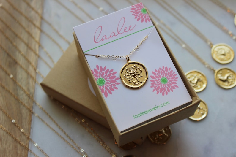 Pu&ograve; includere: Una collana d'oro con un ciondolo rotondo che presenta un design a forma di fiore di loto. La collana &egrave; confezionata in una scatola marrone con un design floreale rosa e verde e il testo "laalee" e "laaleejewelry.com".