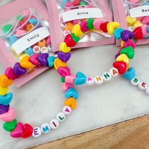 May include: Three colorful heart-shaped bead bracelets with the names "Emily", "Anna", and "Beth" spelled out in white letters. The bracelets are packaged in individual pink bags with the names printed on the front.