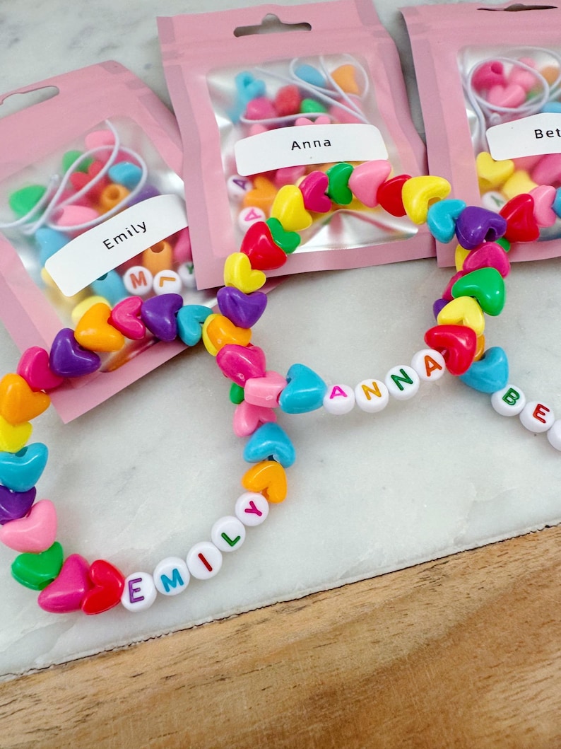 May include: Three colorful heart-shaped bead bracelets with the names "Emily", "Anna", and "Beth" spelled out in white letters. The bracelets are packaged in individual pink bags with the names printed on the front.