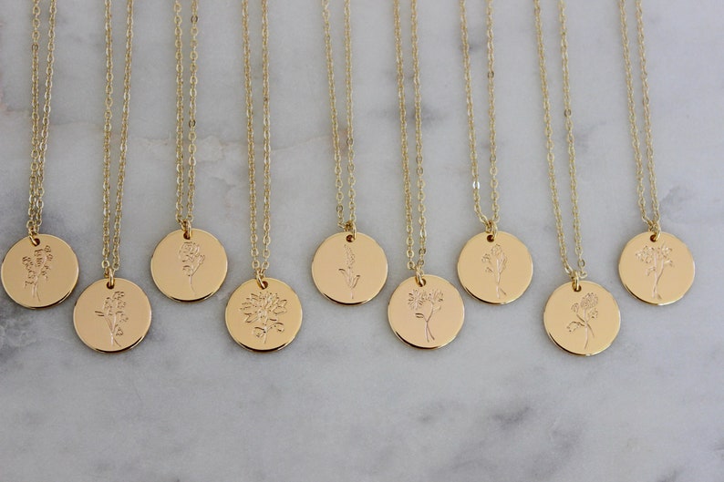 Peut inclure: Colliers dor&eacute;s avec des pendentifs ronds, chacun grav&eacute; d'un motif floral diff&eacute;rent. Les pendentifs sont suspendus &agrave; de fines cha&icirc;nes, cr&eacute;ant une esth&eacute;tique minimaliste et &eacute;l&eacute;gante. Les colliers sont expos&eacute;s sur une surface en marbre.