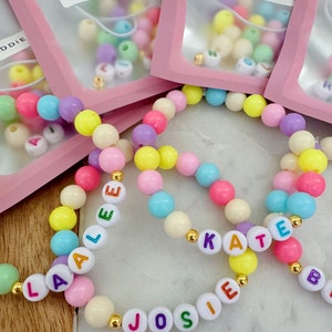 May include: Three colorful beaded bracelets with the names "Laalee", "Josie", and "Kate" spelled out in colorful letters. The bracelets are made with pastel-colored beads and gold spacers.
