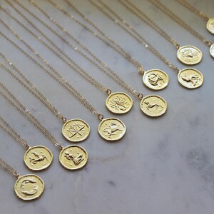 Pu&ograve; includere: Un set di 12 collane zodiacali in oro, ciascuna con un ciondolo con un segno zodiacale diverso. Le collane sono disposte su una superficie di marmo bianco.