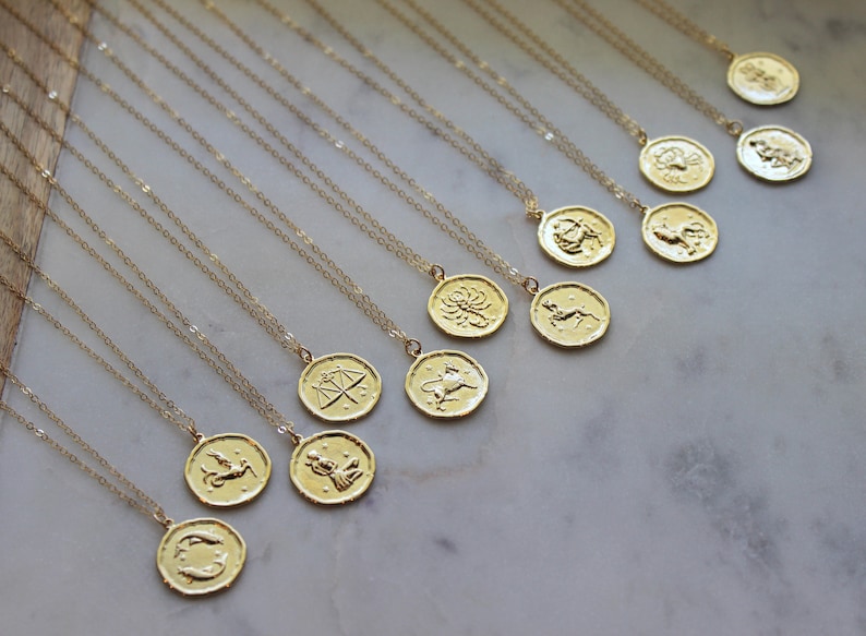 Pu&ograve; includere: Un set di 12 collane zodiacali in oro, ciascuna con un ciondolo con un segno zodiacale diverso. Le collane sono disposte su una superficie di marmo bianco.