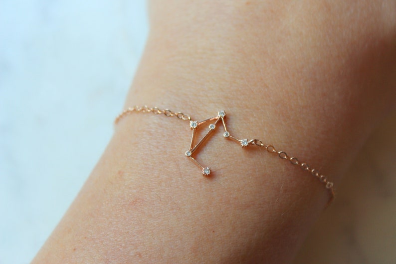 K&ouml;nnte beinhalten: Ein zartes goldenes Armband mit einem Konstellationsdesign, das mit kleinen, klaren Steinen besetzt ist. Die Konstellation ist auf einem Kettenarmband angebracht.