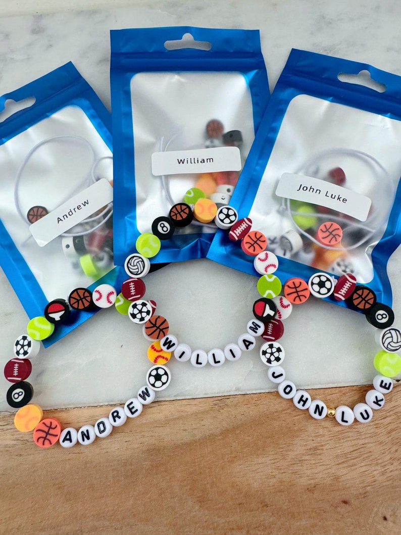 May include: Three clear plastic bags with blue borders and white labels. Each bag contains a set of colorful beads with sports-themed designs and a white label with a name: Andrew, William, and John Luke.