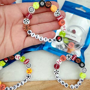 May include: Three bracelets with white beads and colorful sports ball charms. The bracelets spell out the names "William", "Andrew", and "John Luke".