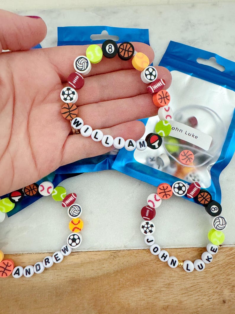 May include: Three bracelets with white beads and colorful sports ball charms. The bracelets spell out the names "William", "Andrew", and "John Luke".