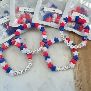 May include: Four red, white, and blue star-shaped bead bracelets with gold lettering spelling out the names "James", "Stella", "Caroline", and "Luke".