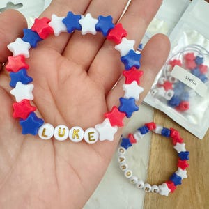 May include: Two bracelets made with red, white, and blue star-shaped beads. One bracelet spells out the name "LUKE" with gold letters on white beads. The other bracelet spells out the name "CAROLINE" with gold letters on white beads.