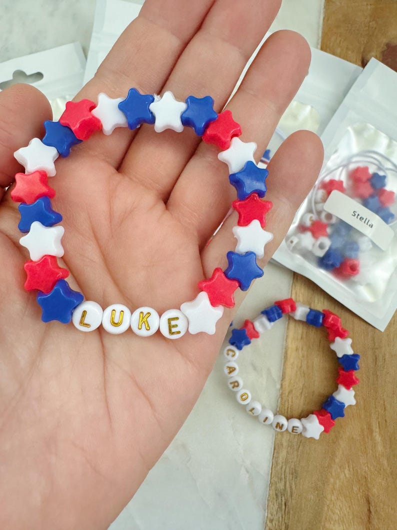 May include: Two bracelets made with red, white, and blue star-shaped beads. One bracelet spells out the name "LUKE" with gold letters on white beads. The other bracelet spells out the name "CAROLINE" with gold letters on white beads.